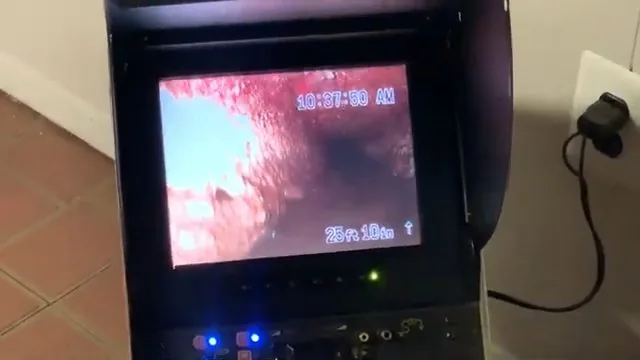 Camera monitor beside a tiled floor showing a red-tinted pipe wall with a distance reading in the corner.