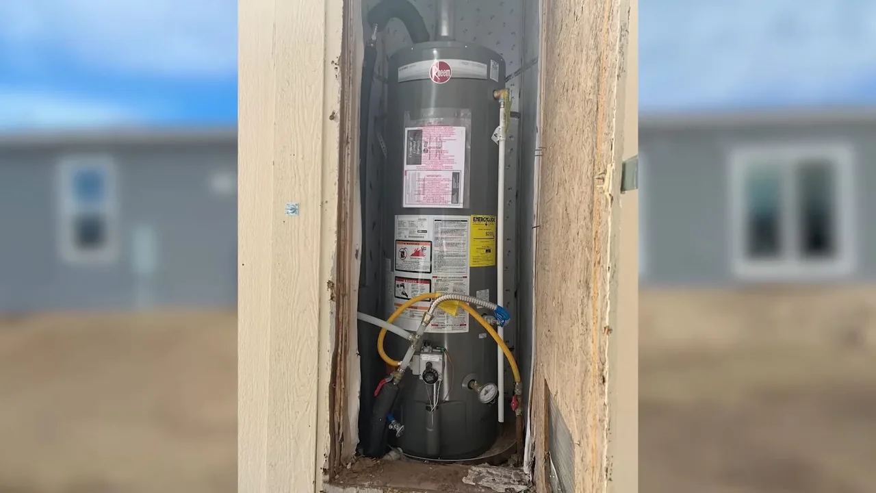 Tank water heater in a narrow framed closet with a yellow gas line, a pale manufactured home visible behind it.