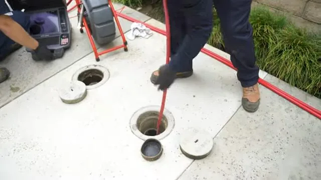 Technician in work boots feeding a red camera cable into a cleanout in paving, with the reel and monitor to the left.