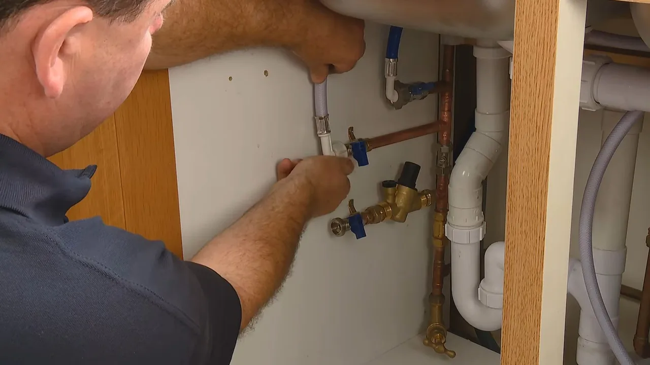 Technician in a dark polo reaching into a sink cabinet toward blue-handled shutoff valves and supply lines.