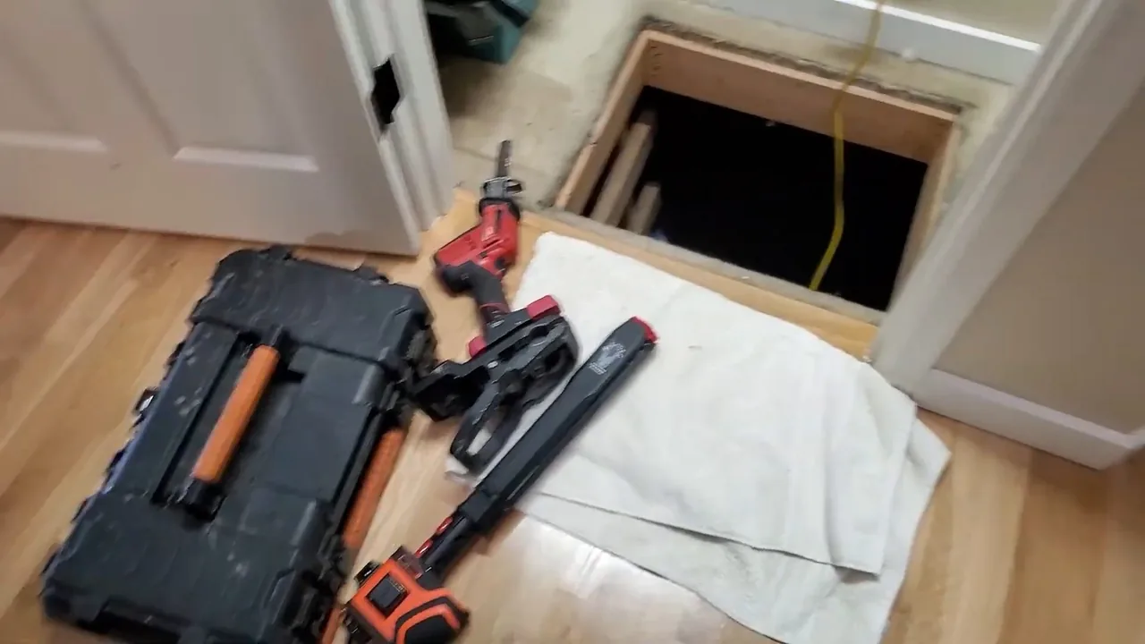 Tool case, power tool and wrench on a towel on a wood floor beside an open floor access hatch.
