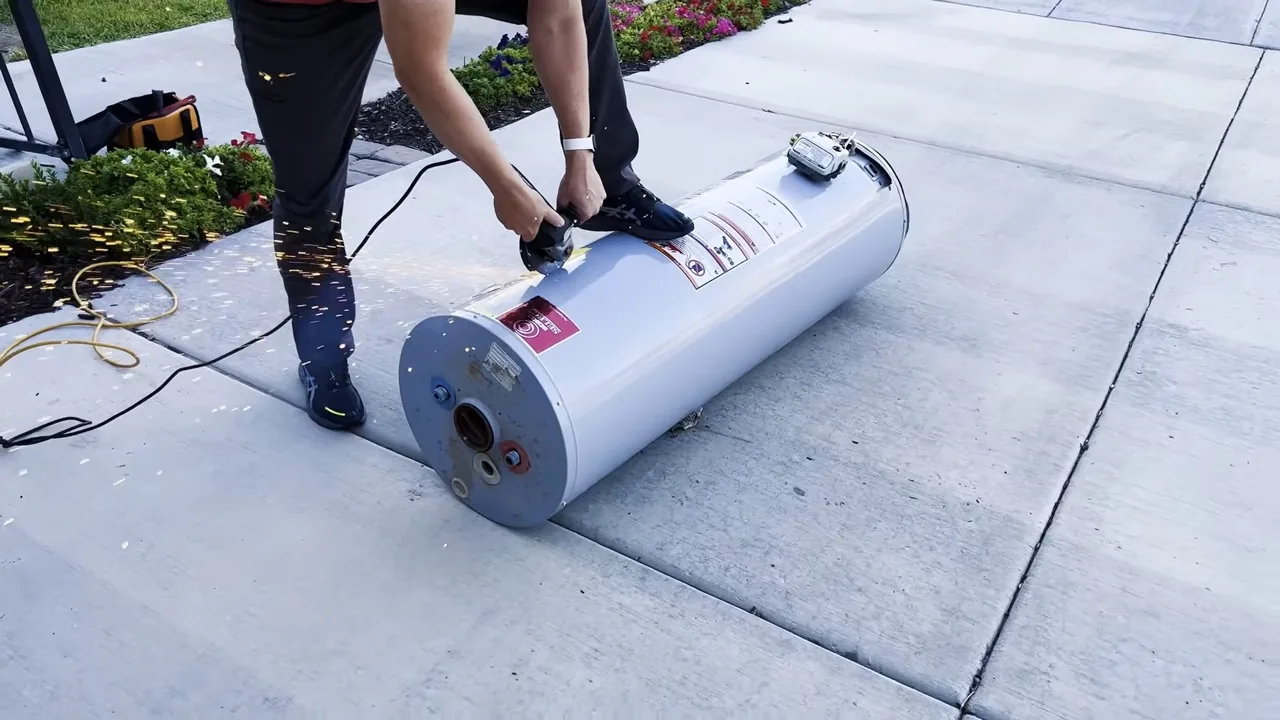Two people on a concrete patio rolling a white tank water heater along on its side.