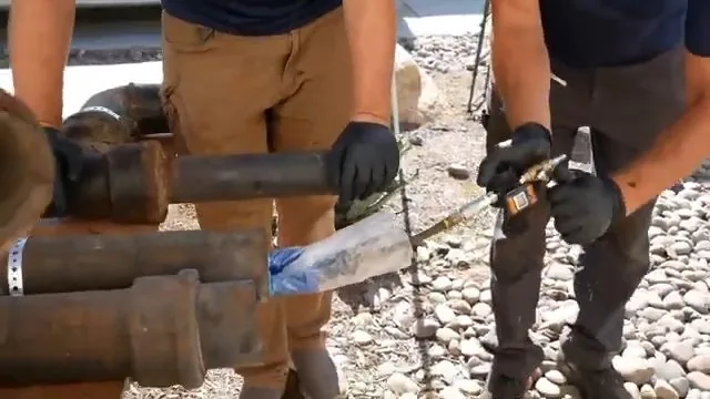 Two workers in gloves standing over short lengths of cast iron pipe on gravel, one feeding a clear sleeve into a pipe end.