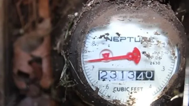 Close view of a dirty water meter dial showing a red sweep hand and the cubic feet readout.