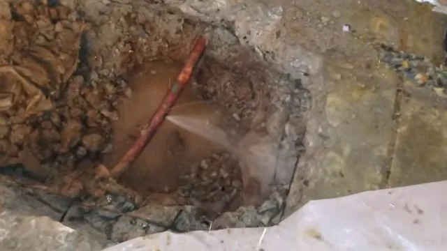 Opening dug through a floor showing a red supply line with water spraying from it into muddy soil.