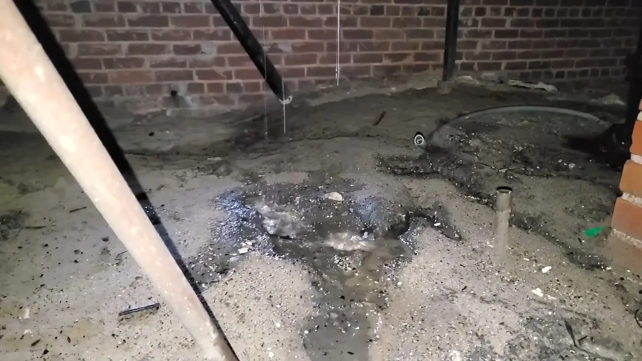 Wet debris and standing water on bare ground beneath a pipe, beside a brick foundation wall.