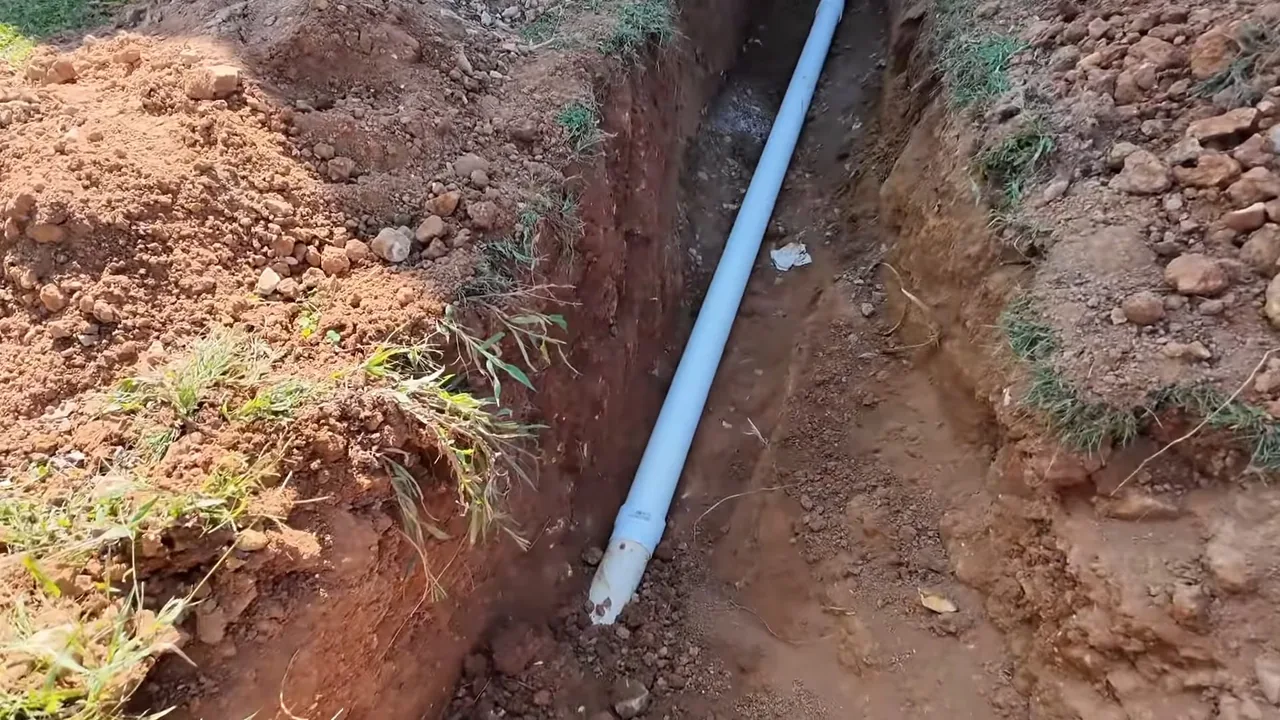 Narrow trench through red-brown soil with a long white pipe lying in the bottom.