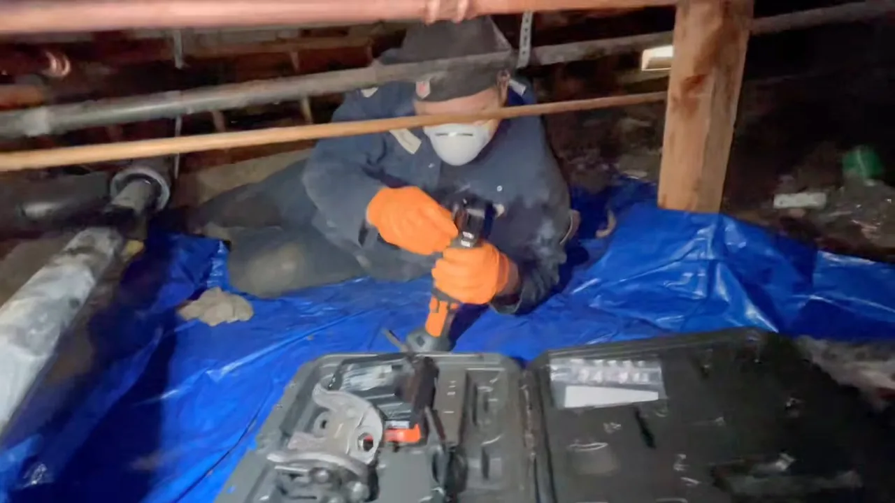 Worker in a dust mask and orange gloves repairing a pipe in a crawlspace, kneeling on a blue tarp beside an open tool case.